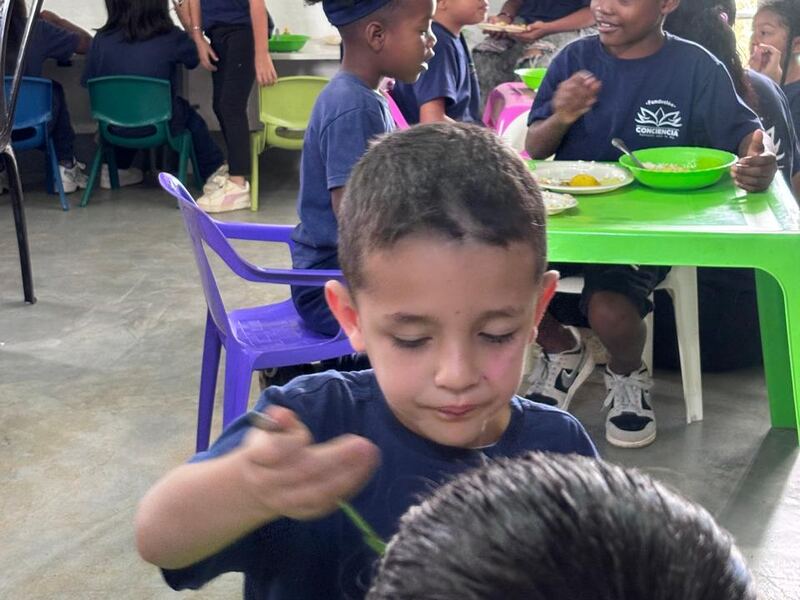 Un niño almuerza en el comedor comunitario de la fundación, con otros niños en mesas de colores al fondo.